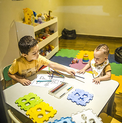 Duas crianças sobre a mesa, brincando com lápis de cor e desenhos para colorir.