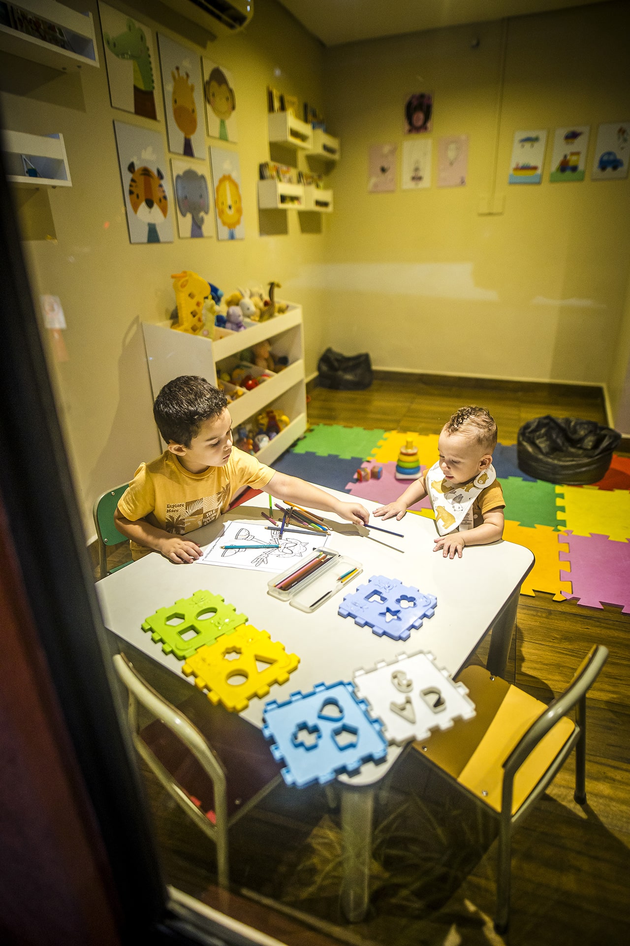 Duas crianças sobre a mesa, brincando com lápis de cor e desenhos para colorir.